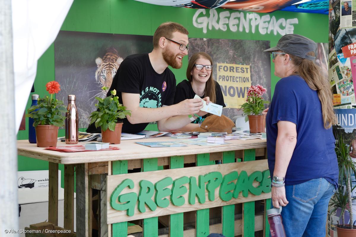 At a Greenpeace branded stall, two smiling volunteers speak to a member of the public.