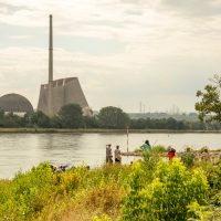 People play in lush vegetation by a river. On the opposite bank sits a large nuclear power station in the process of being demolished.