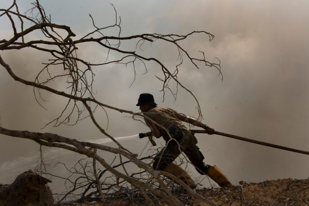 A firefighter strains forward to pull a spraying hose closer to a forest fire. Grey-brown smoke fills the background.