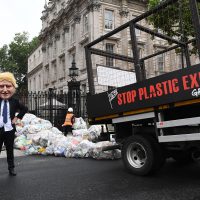 A tipper truck with 'Stop plastic exports' stencilled on the side drops bags of plastic waste outside the gates of Downing Street. An activist wearing a caricatured Boris Johnson mask looks on.