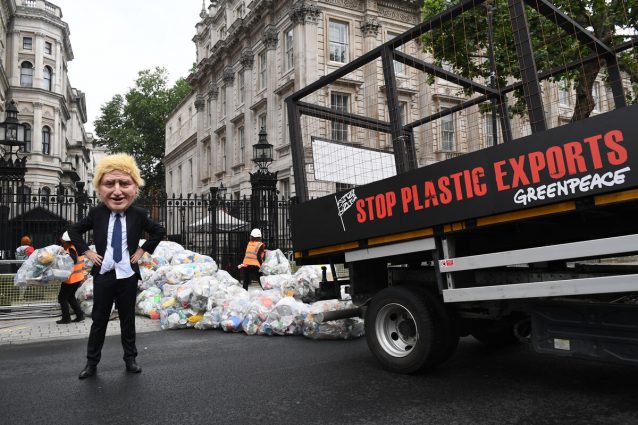 A tipper truck with 'Stop plastic exports' stencilled on the side drops bags of plastic waste outside the gates of Downing Street. An activist wearing a caricatured Boris Johnson mask looks on.