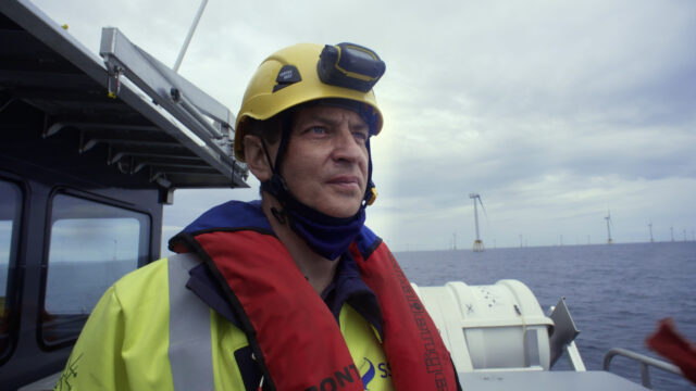 Alan (hard hat, life jacket, hi-vis) looks out on an offshore windfarm from the deck of a work boat