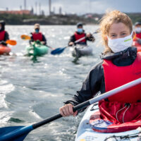 An activist (red buoyancy aid, long hair tied back, cloth facemask) smiles into the camera. A row of other kayakers is visible in the background.