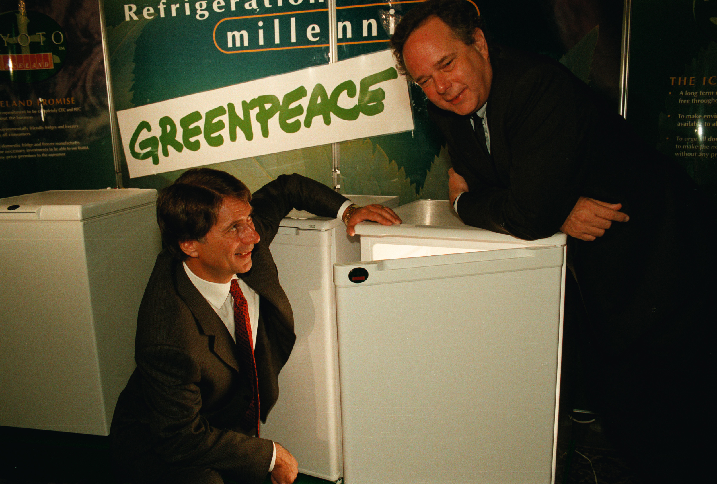 Two suited men stand around a half open freezer with a banner reading Greenpeace in the background