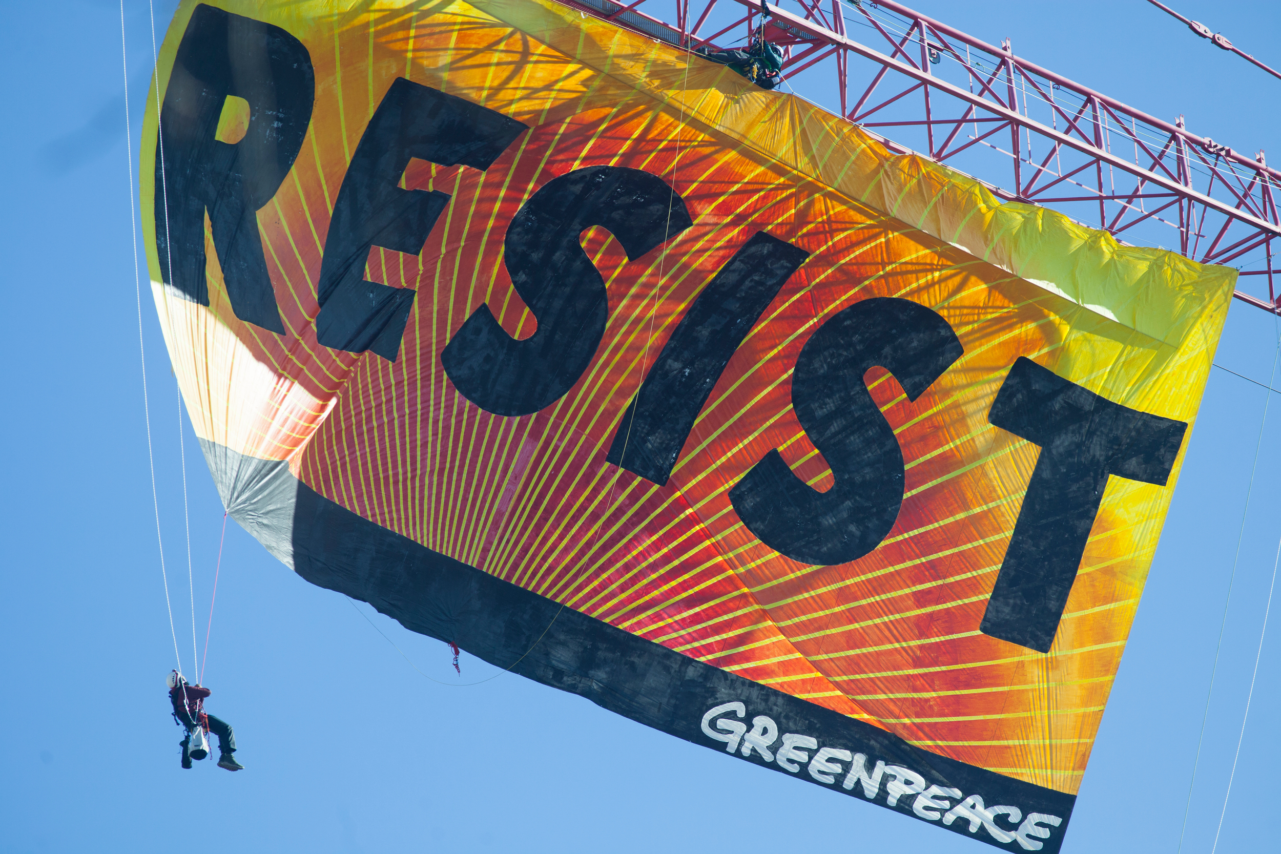 a huge orange and yellow banner filling the frame hanging from a crane reading RESIST with a person dangling off the left bottom corner