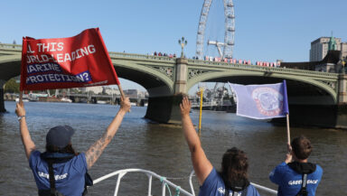 Three Greenpeace crew members wave and hold up banners towards a group of protesters on a bridge. The protesters are holding up placards reading 'No fish no future'. The 'London Eye' ferris wheel is visible in the background.