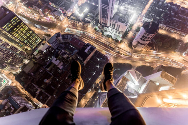 First-person view of someone sitting on the edge of a skyscraper at night. The photographer's legs and feet are seen hanging over the edge, and tall buildings and streets are visible far below.