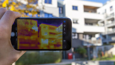 A thermal imaging camera held up to a low-rise apartment building. The camera's screen shows the temperature of different parts of the building's exterior, colour-coded in yellow, orange and purple.