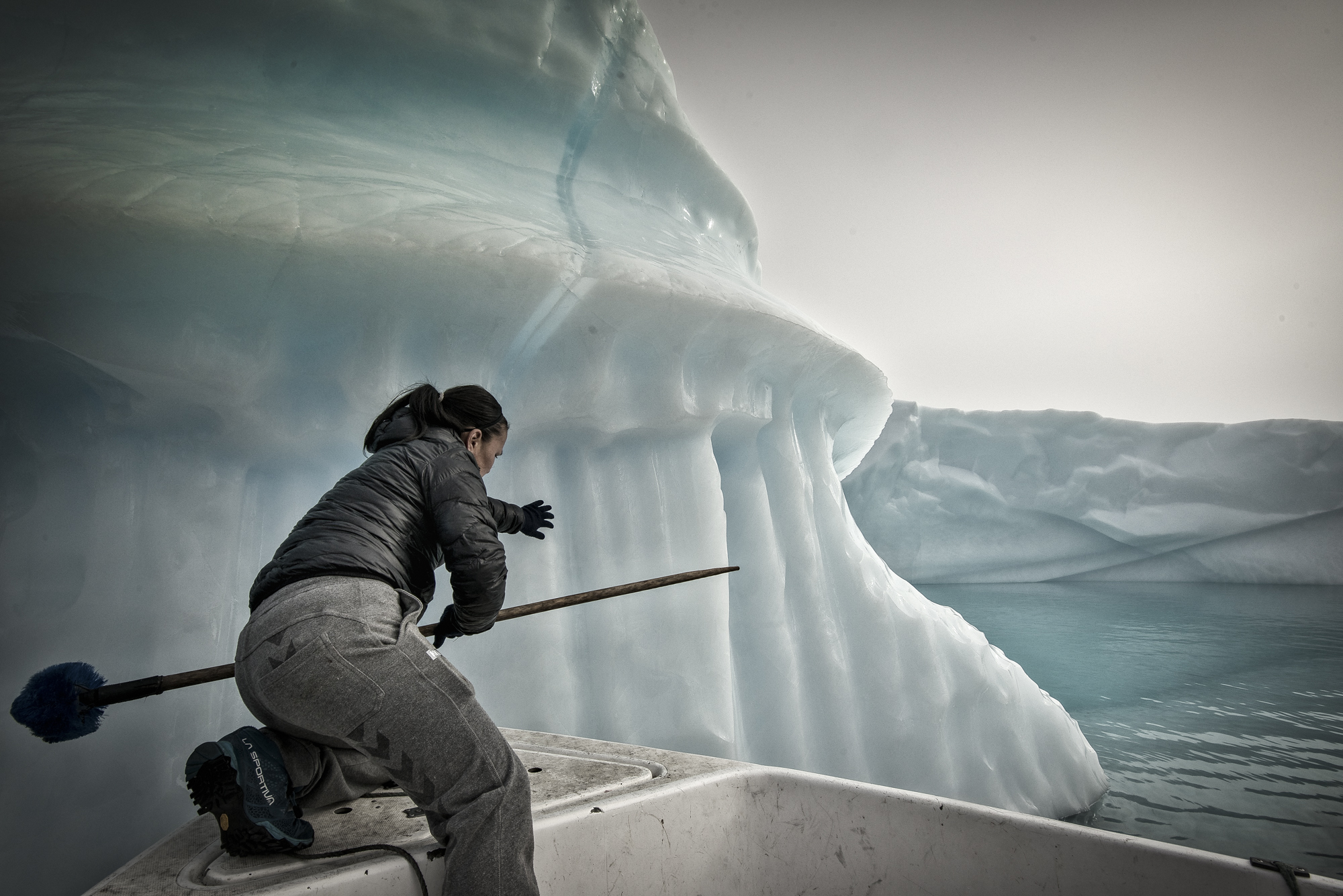 A person in jeans and a puffer jacket carrying an oar, at the front of a small boat, pushing an iceberg that's so big the top is out of shot, in a pale-blue icy seascape