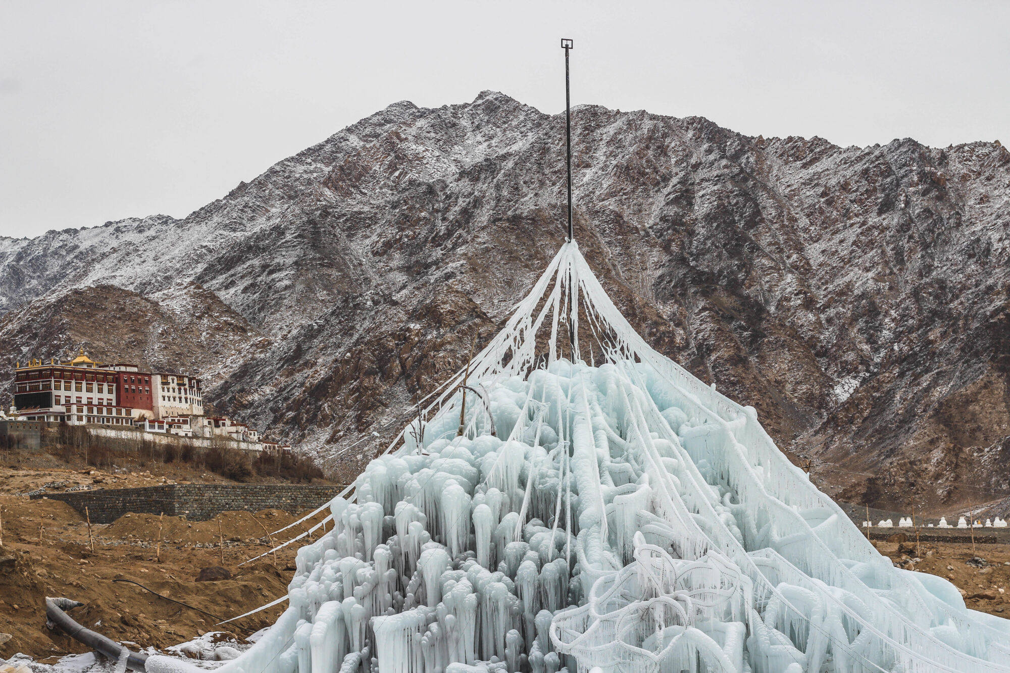 An early prototype of artificial glacier, named Ice Stupa - the device appears like a sculpture, with long lines of icicles rising up to a central pole. Beneath this large clusters of ice form in unusual shapes, some spirals of ice, others large clumps of ice with thick icicles dripping down.