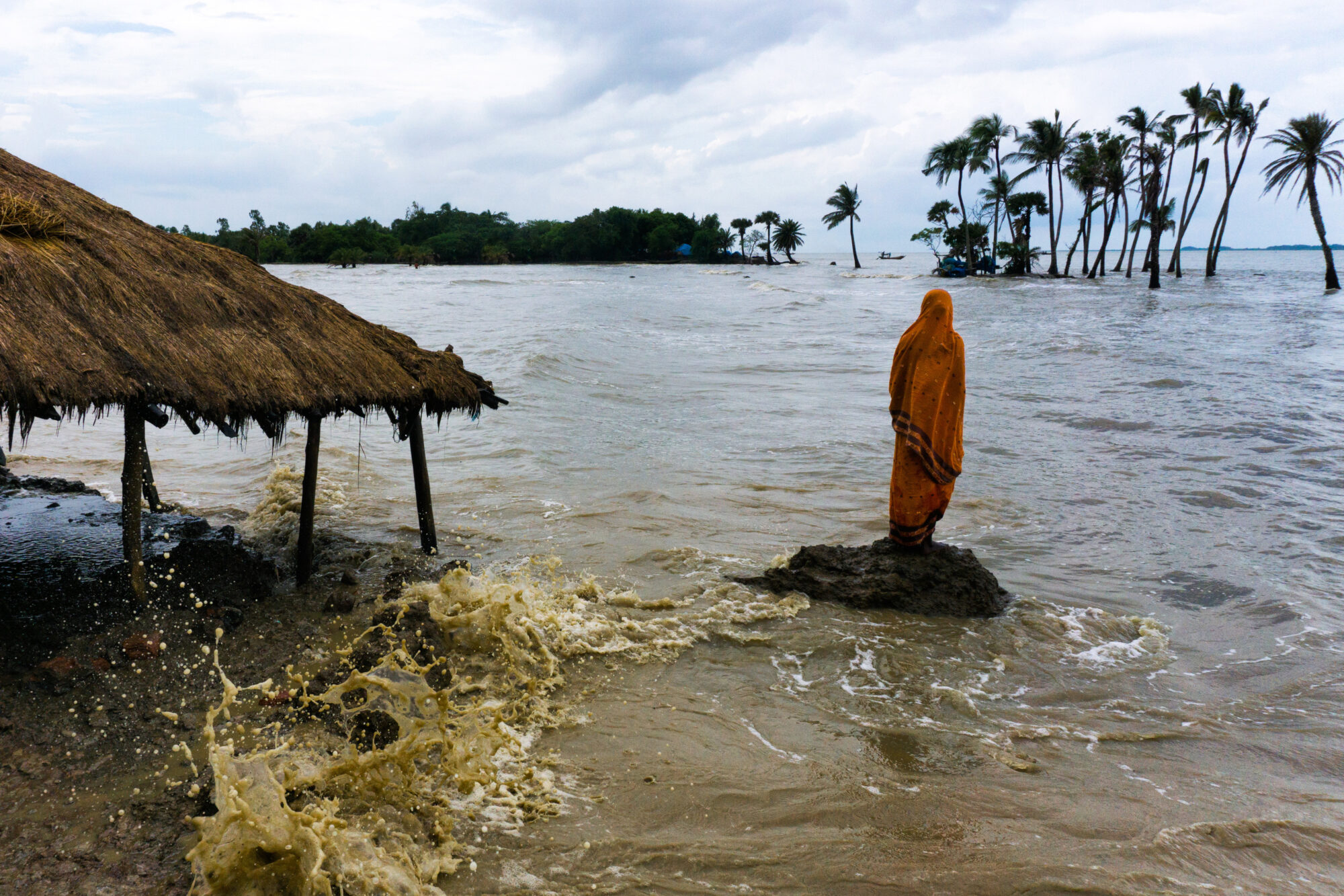 A person wearing traditional sari stands on a rock, surrounded on all sides by rising dirty sea water. The waves of brown water break against a small hut to the left.