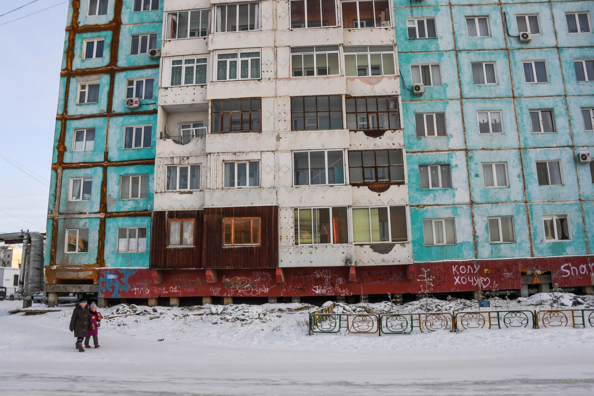 Two people walk along a snowy street in front of a large apartment building. The building's paint is fading, panels are cracked, and is covered in graffiti.
