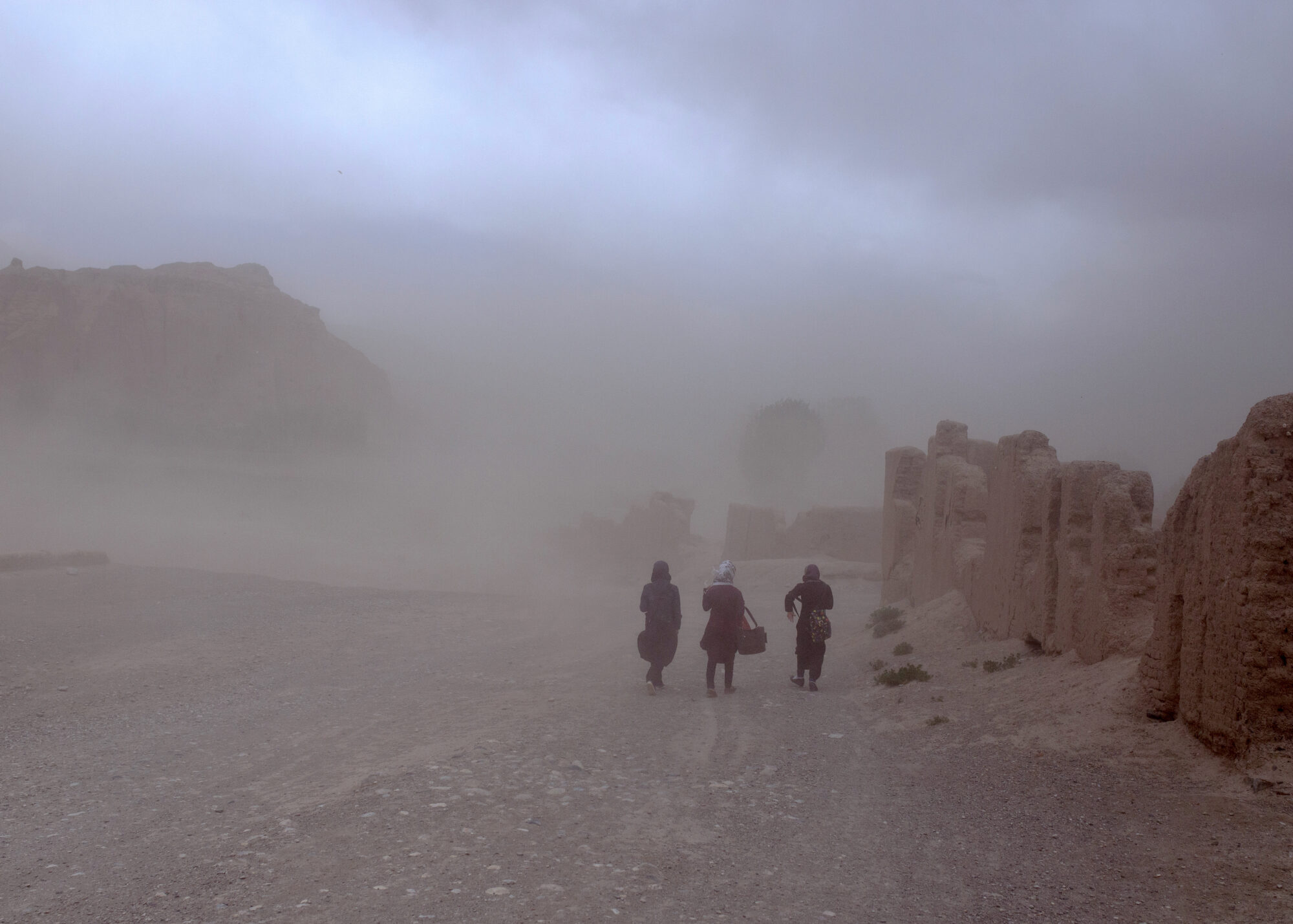 Three people walk along a dirt road past stone ruins. A thick layer of dust hangs in the air, obscuring the view of the distance.