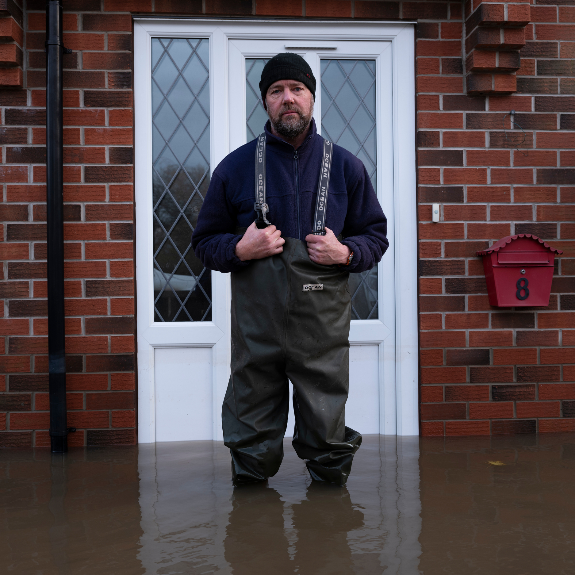 A person wearing fishing trousers standing in front of a red brick house, with dark flood water up to their knees.