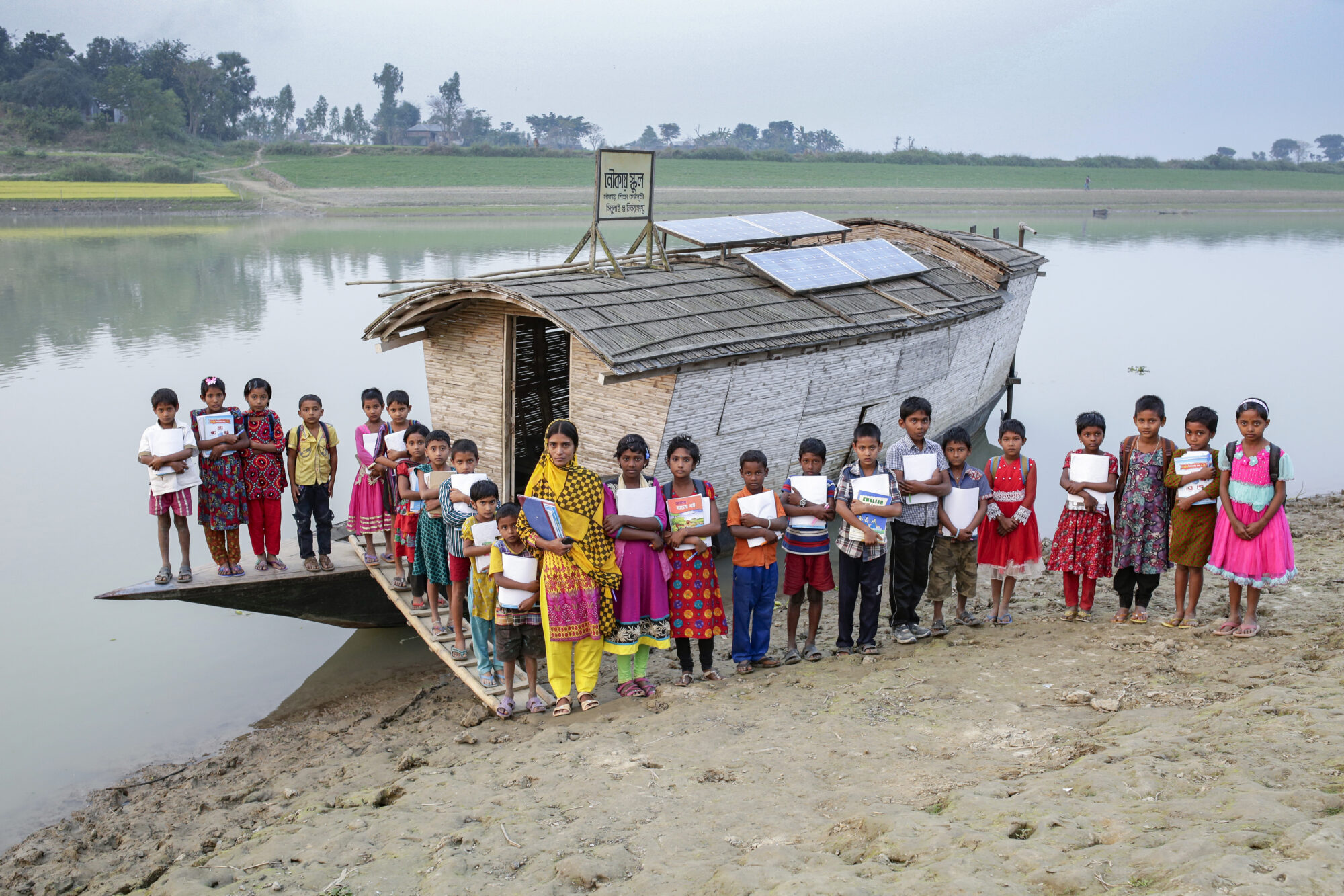A group of school children and their teacher stand together outside of a small boat with a wicker roof on the banks of a river.