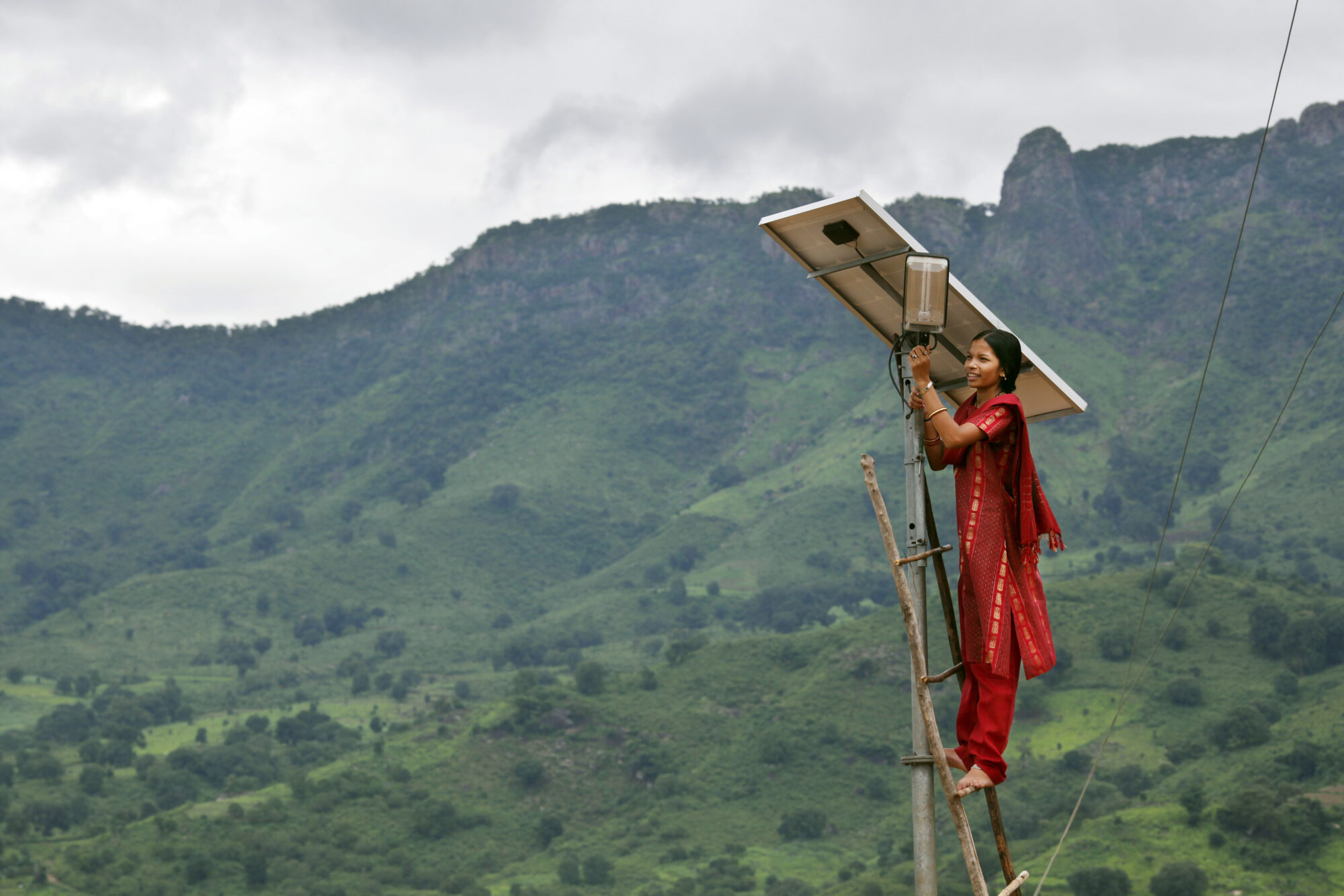 A person stands barefoot at the top of a tall ladder, making adjustments to the mounted solar panel. Rolling mountains seen behind them, giving a sense of the tall height they are working at.