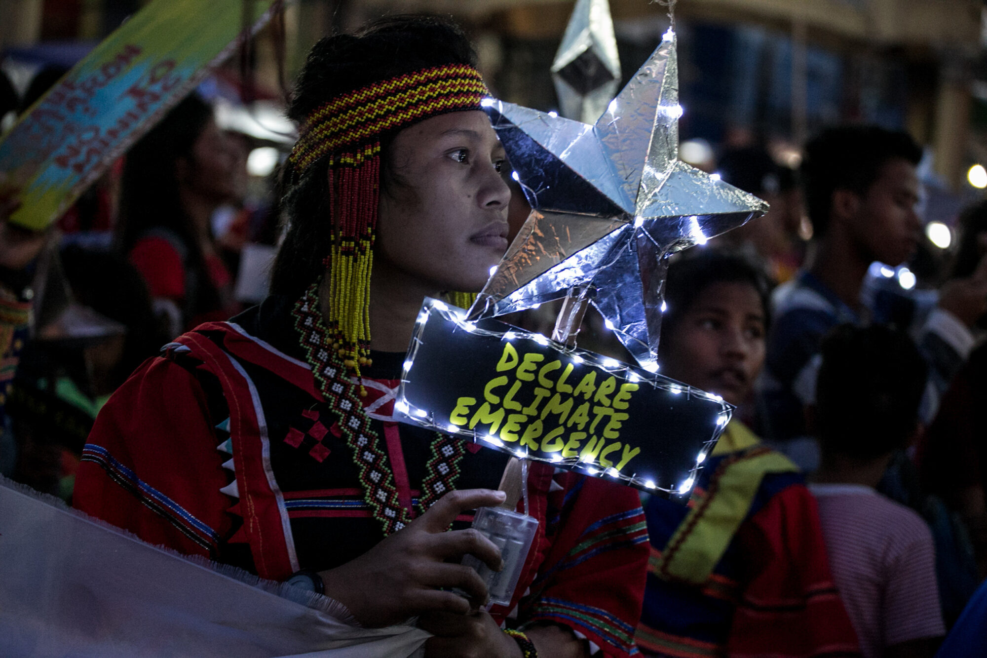 A climate activist carrying a placard in the shape of a star, with fairy lights around it, and sign reading "Declare Climate Emergency".