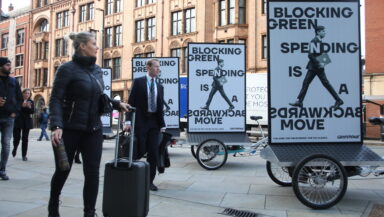 Conference delegates in business suits walk past a trio of cargo bikes with billboards mounted on the back. Each one shows a photo of Rishi Sunak photoshopped so his legs are facing backwards. Text reads 'Blocking green spending is a backwards move'.
