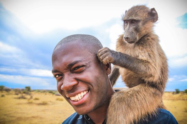Patrick Aryee with a monkey sitting on his shoulder pulling his ear. He's closing one eye and laughing and looking towards the camera