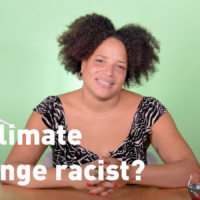 A video presenter (dark curly hair, light brown skin, black and white top) sits at a table decorated with models of a wind turbine and Greenpeace ship. She smiles into the camera. Overlaid text reads 'Is climate change racist?'