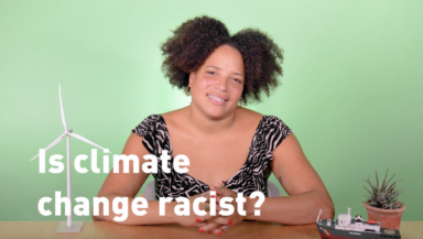 A video presenter (dark curly hair, light brown skin, black and white top) sits at a table decorated with models of a wind turbine and Greenpeace ship. She smiles into the camera. Overlaid text reads 'Is climate change racist?'