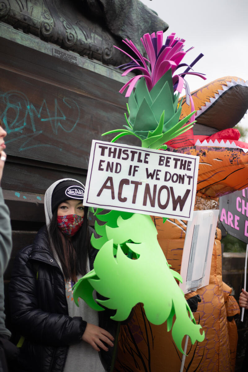 A climate marcher holds a giant paper model of a thistle, with a sign saying 'Thistle be the end if we don't act now'