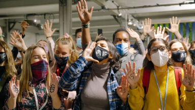 A group of masked young activists gathered round the camera. They're energetic and animated, with hands raised or cupped to their mouths as they chant slogans.