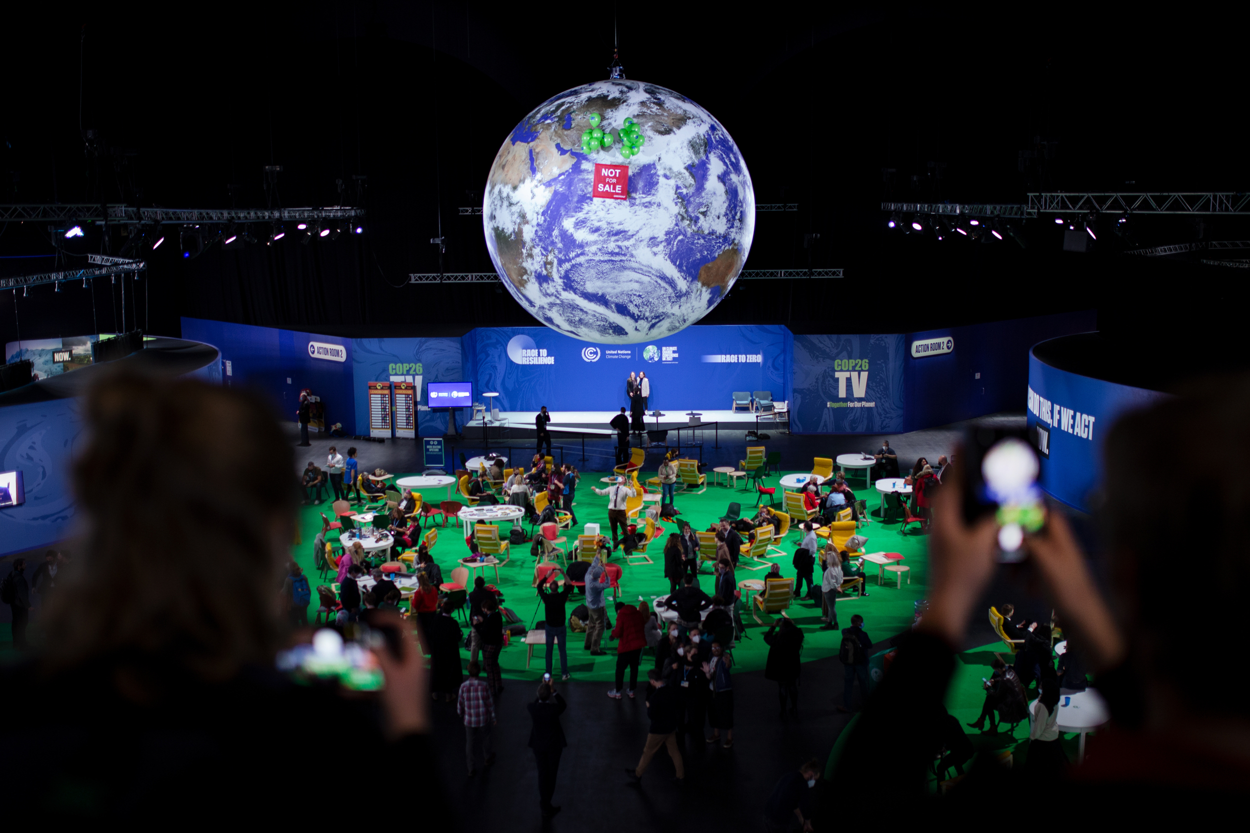 Wide view of the central concourse at COP26. People are gathered underneath a giant globe that hangs overhead. Lifted by green balloons, a banner floats in front of the globe reading 'Not for sale'.