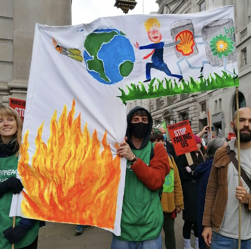 Climate marchers carry a painted banner showing Boris Johnson, plus Shell and BP oil barrels with legs, pushing the Earth off a cliff into a lake of fire.