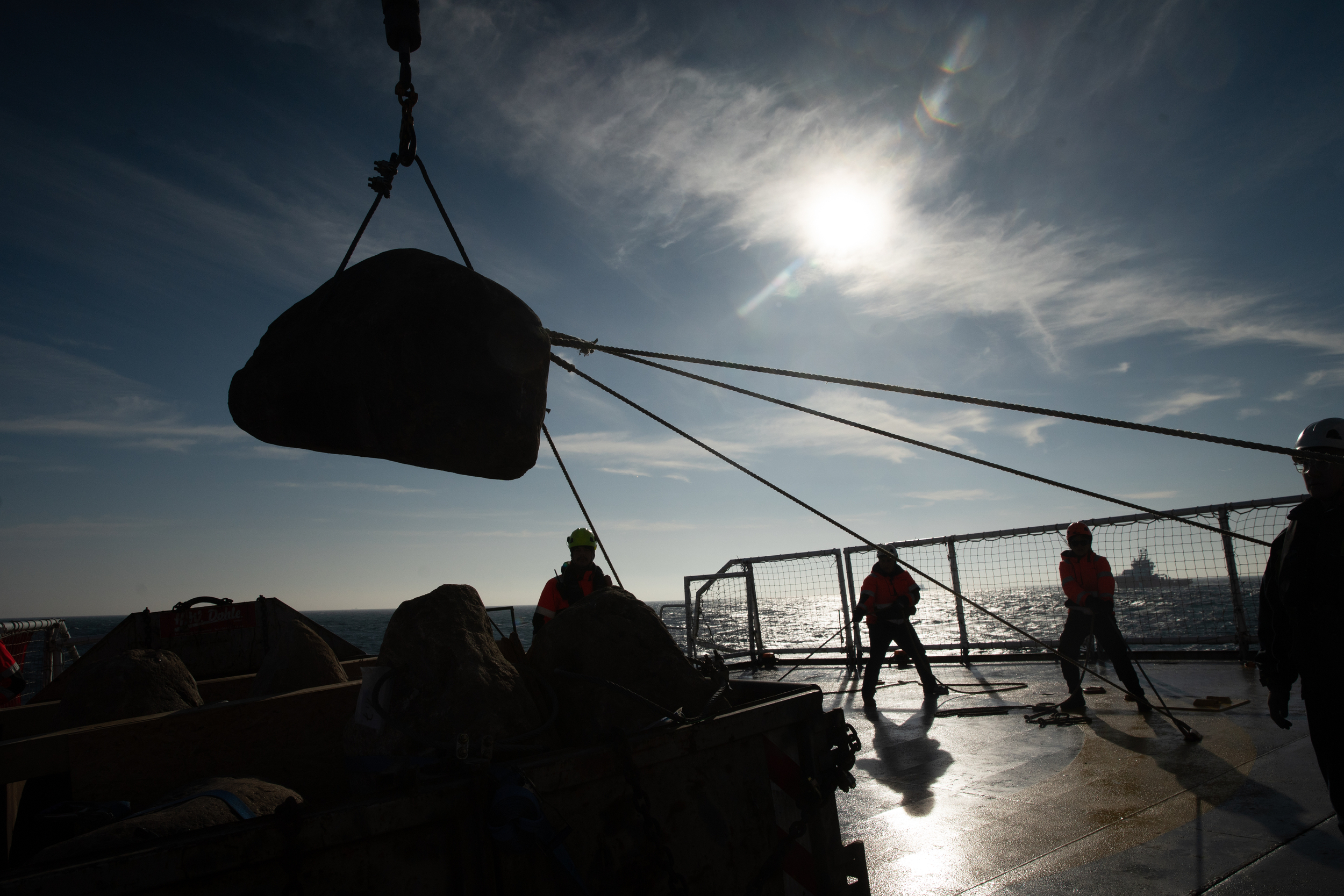 A boulder is silhouetted against a bright blue sky as it's lifted off the deck of a ship by a crane. Crew members surround the boulder, using ropes to control its movement.