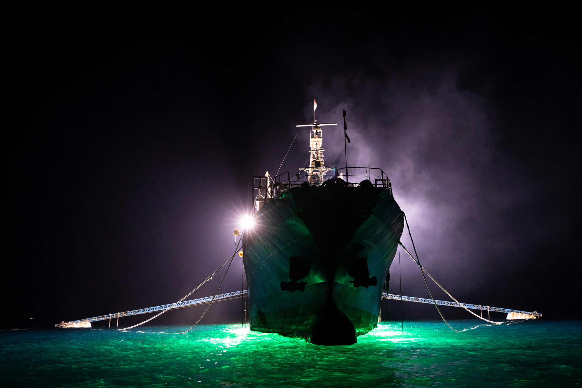 Photographed from below, the prow of a giant fishing boat looms out of the darkness, surrounded by a halo of mist. Bright lights used to attract squid make the surrounding water appear bright green.