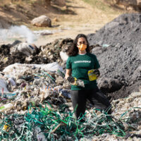 A masked investigator in a Greenpeace tshirt stands in a desolate-looking waste dump. Smoke rises from the piles of smouldering plastic in the background. She holds up a yellow piece of plastic packaging with English writing visible on the front.