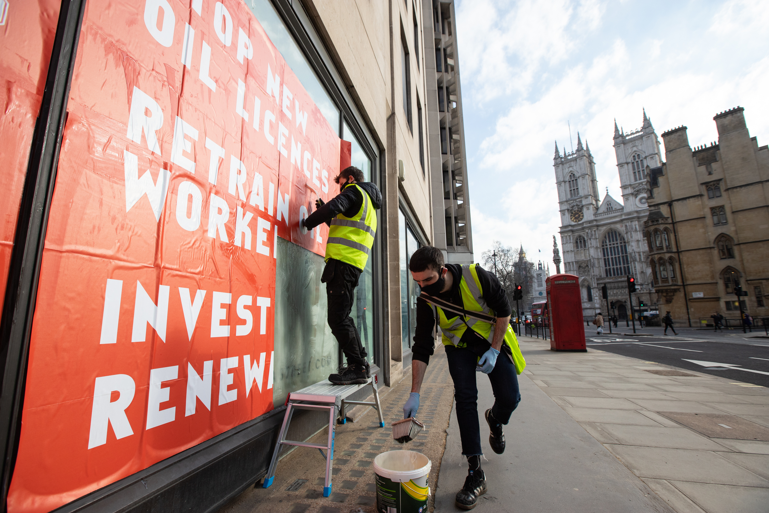 Activists in hi-vis jackets paste a large poster onto a window in a city street. A cathedral is visible in the background. Slogans on the poster say 'Stop new oil licenses. Retraining for workers. Invest in renewables'.