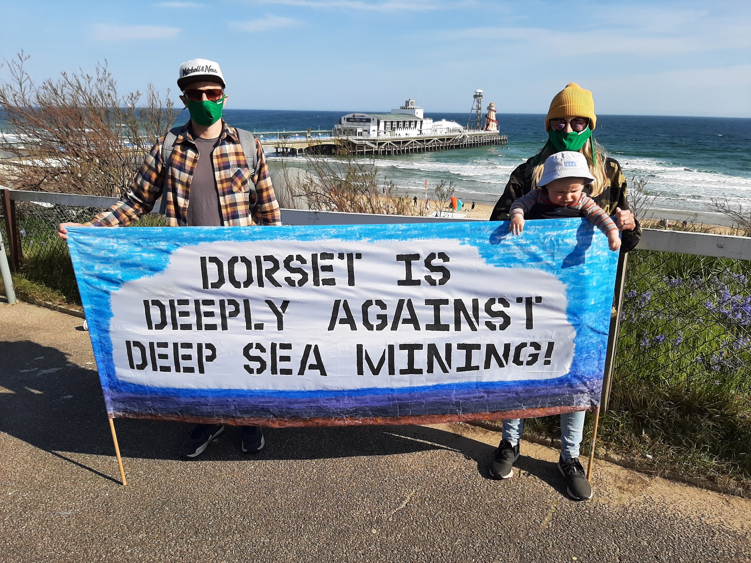Two activists in green masks stand behind a large banner reading 'Dorset is deeply against deep sea mining'. The activist on the right carries a baby in a front-mounted carrier, and in the background a Victorian pier reaches into a calm blue sea.