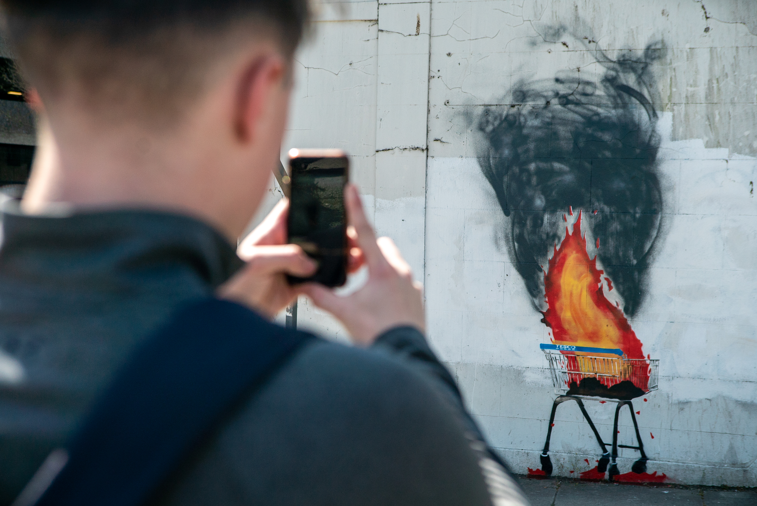 View over the shoulder of a member of the public taking a photo of a mural showing a burning supermarket trolley pouring with black smoke.