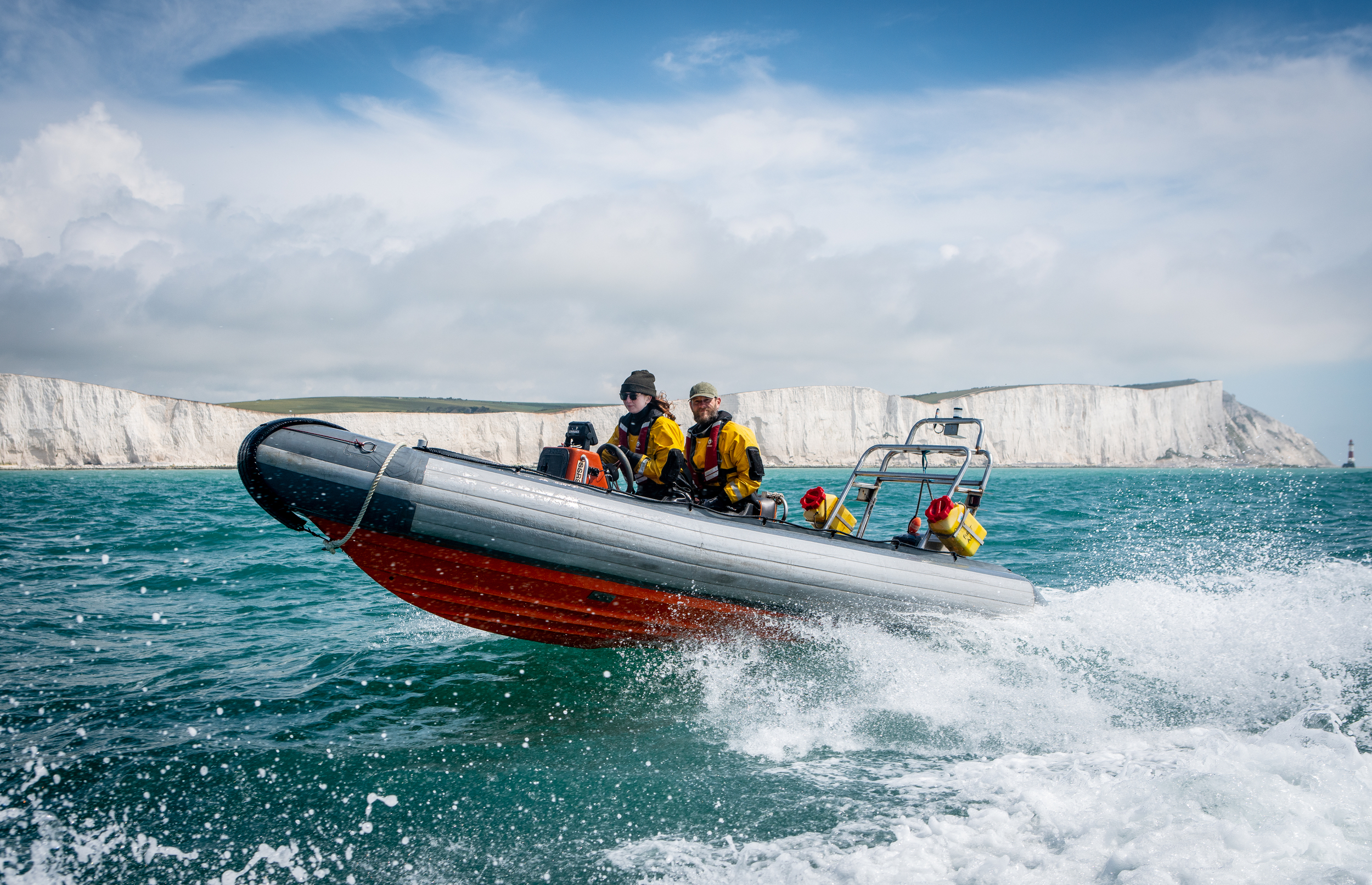A rigid-hulled inflatable boat speeds through the water with activists in yellow survival suits on board. Tall white chalk cliffs are visible in the background.