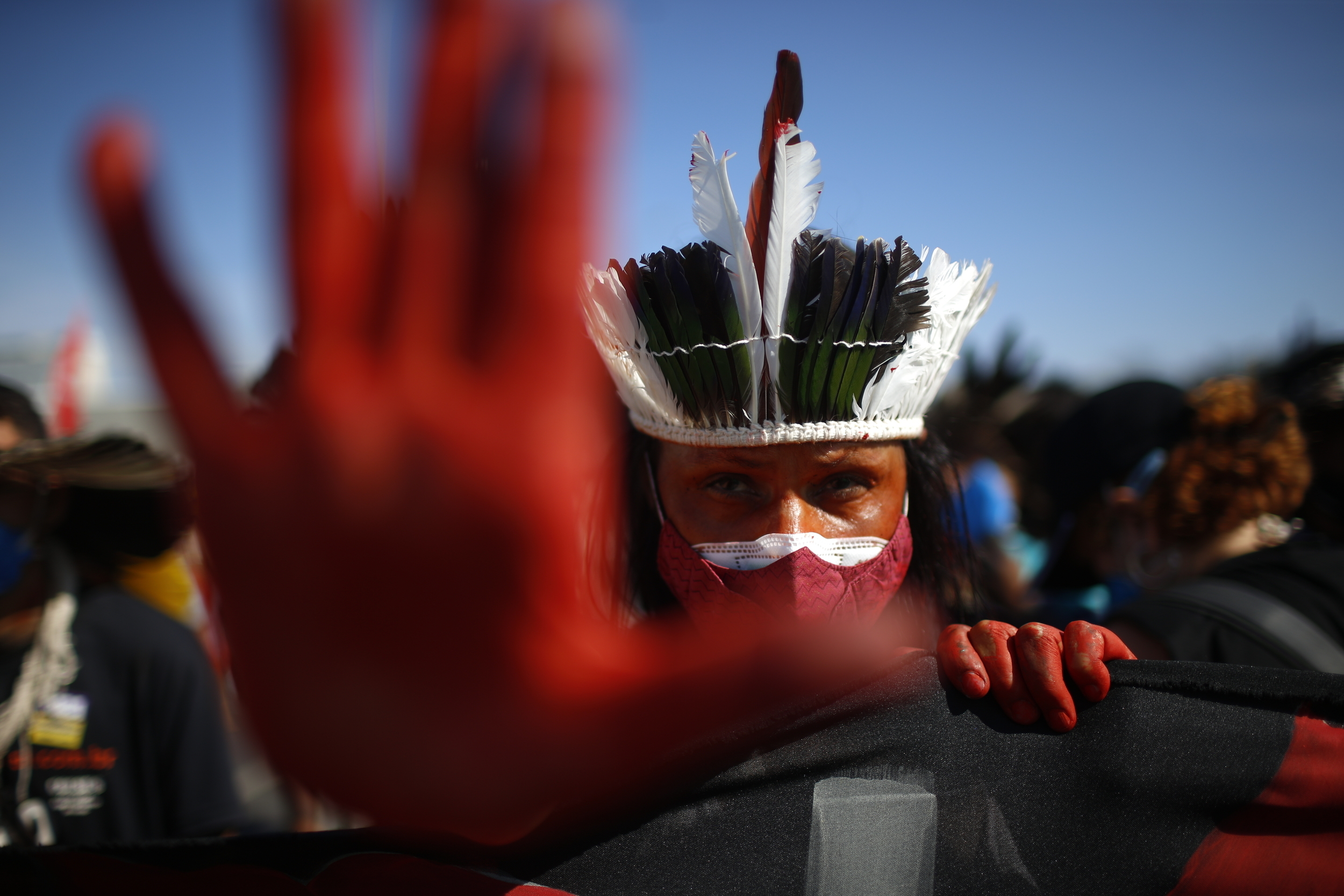 An Indigenous person in a traditional feather headdress and face paint holds a red-painted open palm up close to the camera.