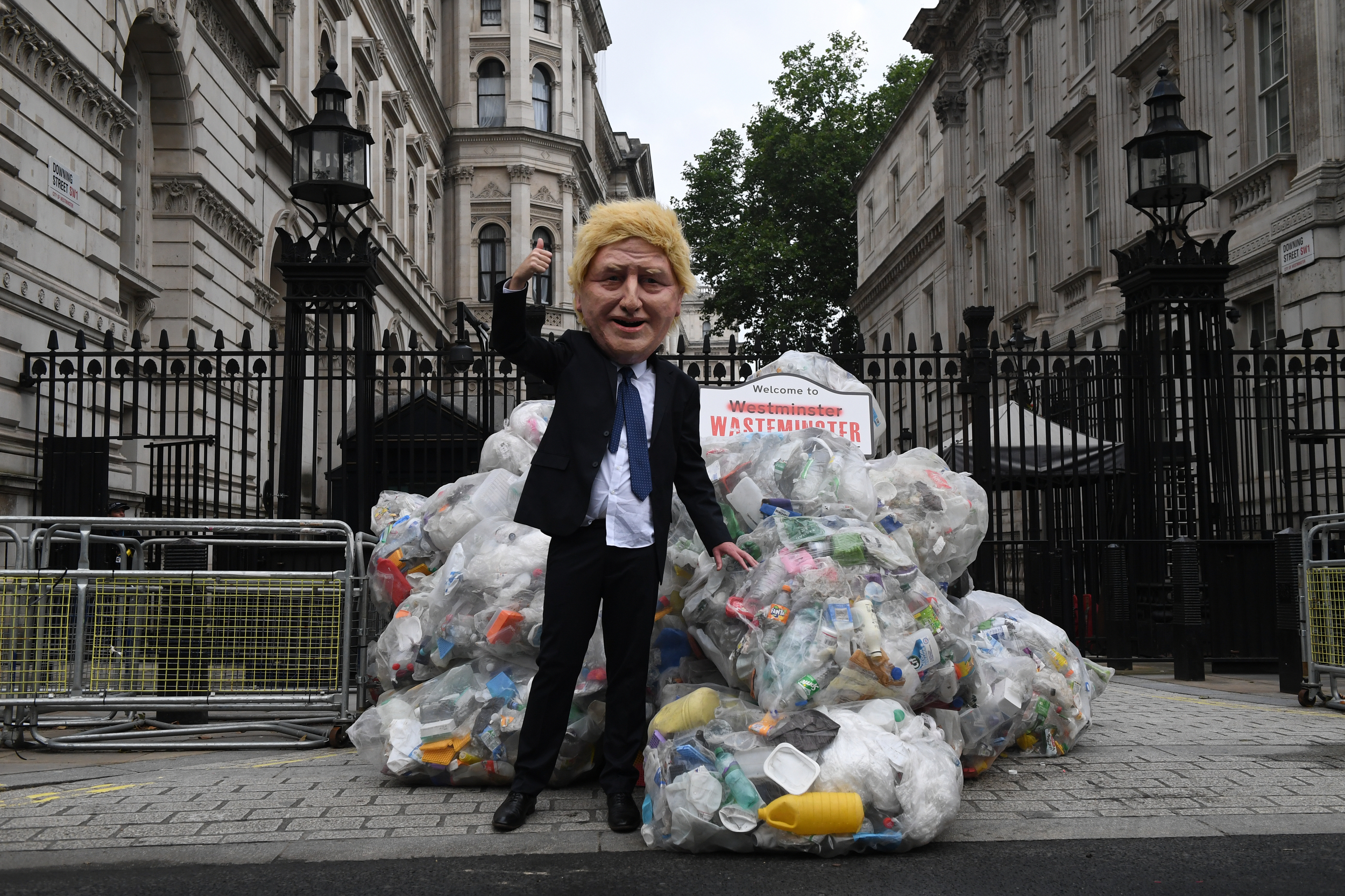 A suited figure in a caricatured Boris Johnson mask stands in front of a huge pile of transparent bin bags filled with plastic waste. In the background, the railings and buildings of Downing Street are visible.