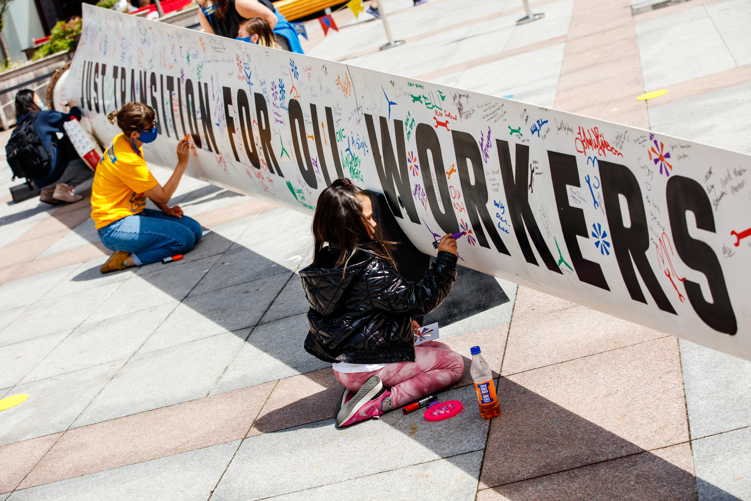 In a sunny city square, three people write messages on a large white wind turbin blade with coloured pens. Large block capitals on the blade read 'Just transition for oil workers'.
