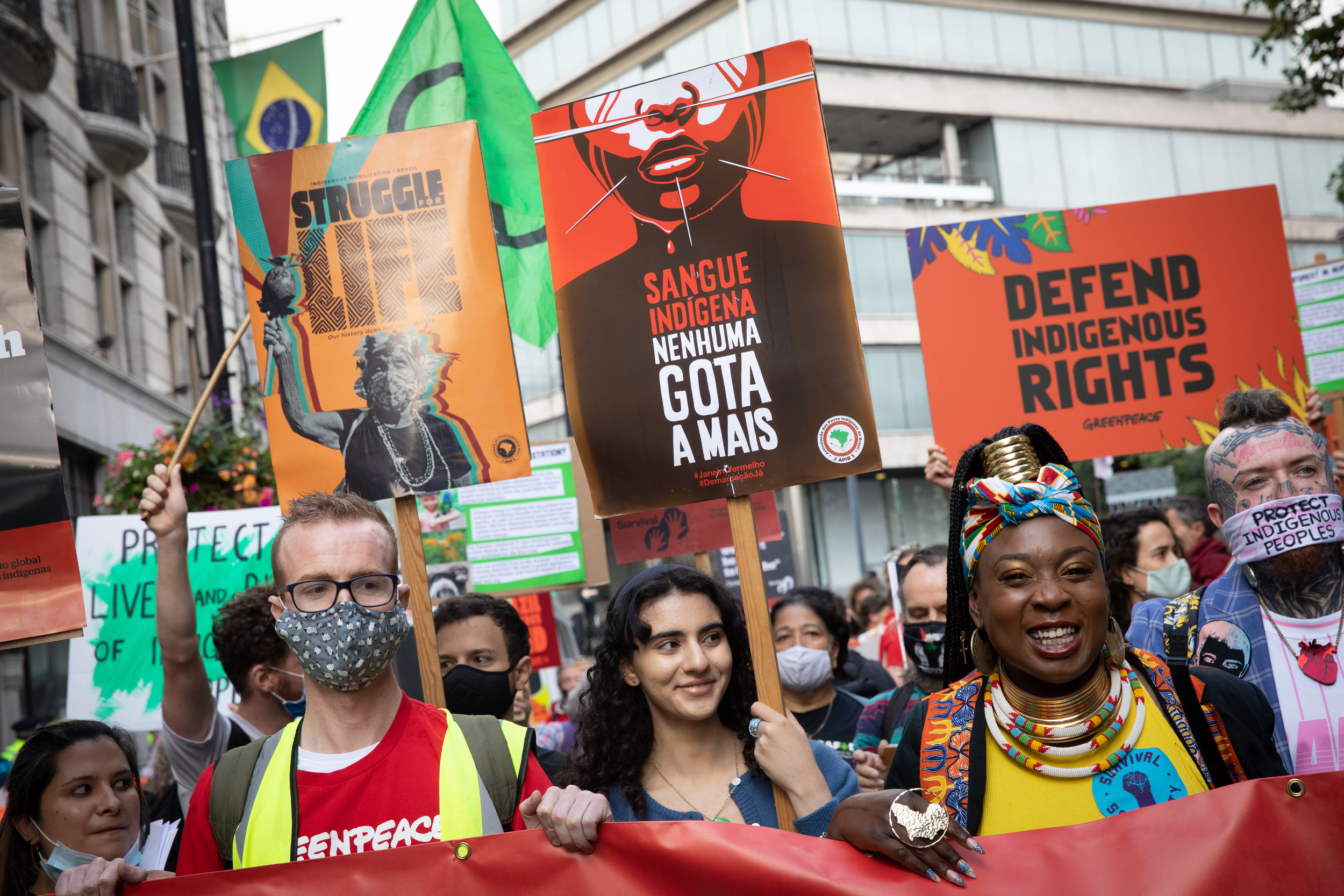 A group of protestors at a march, smiling. They hold banners, which read "Struggle [for] life", "Defend Indigenous rights", "protect indigenous peoples". One sign in Portugese, reads "Sangue Indigena Nunhuma Gota a mais"