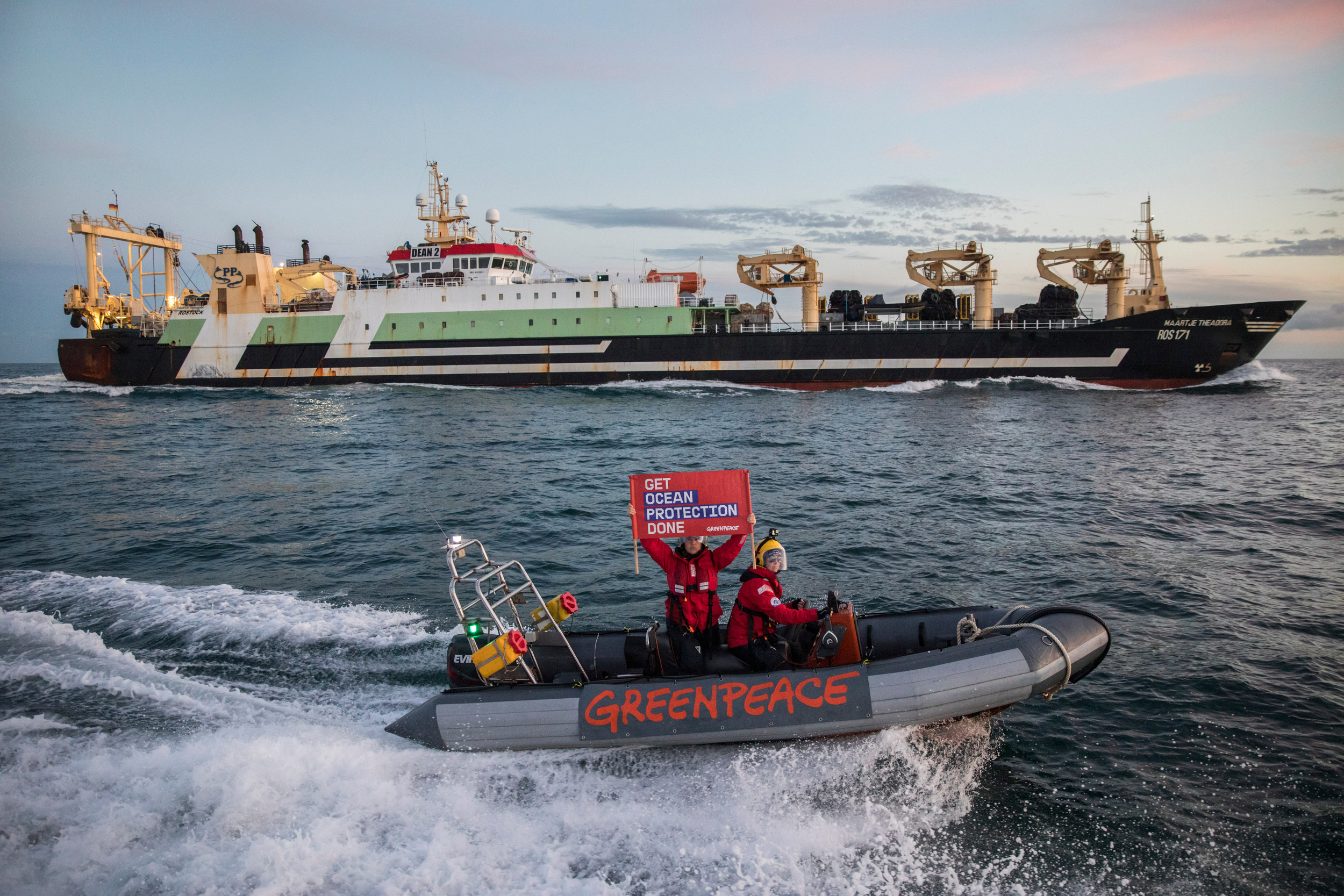 Two people on a speed boat, with "Greenpeace" on the side, sail past a huge industrial fishing ship. One person in the speedboat holds a banner saying "Get Ocean Protection Done".