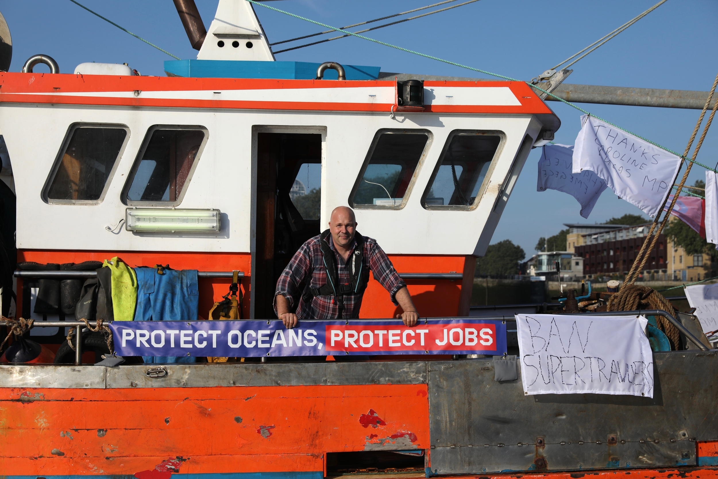 A fisherman leans over the rail of his small boat, smiling at the camera. Banners hang from the side of the boat, reading "Protect oceans, protect jobs" and "Ban Supertrawlers".