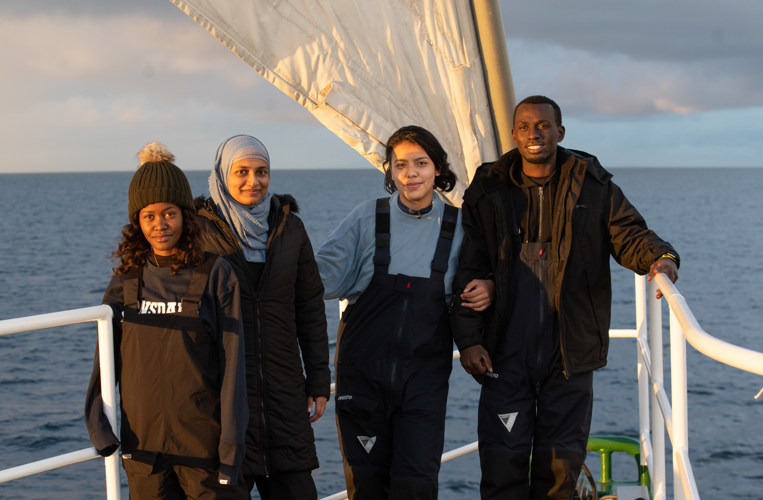 Four young activists stand on the deck of a ship in warm evening light, smiling at the camera