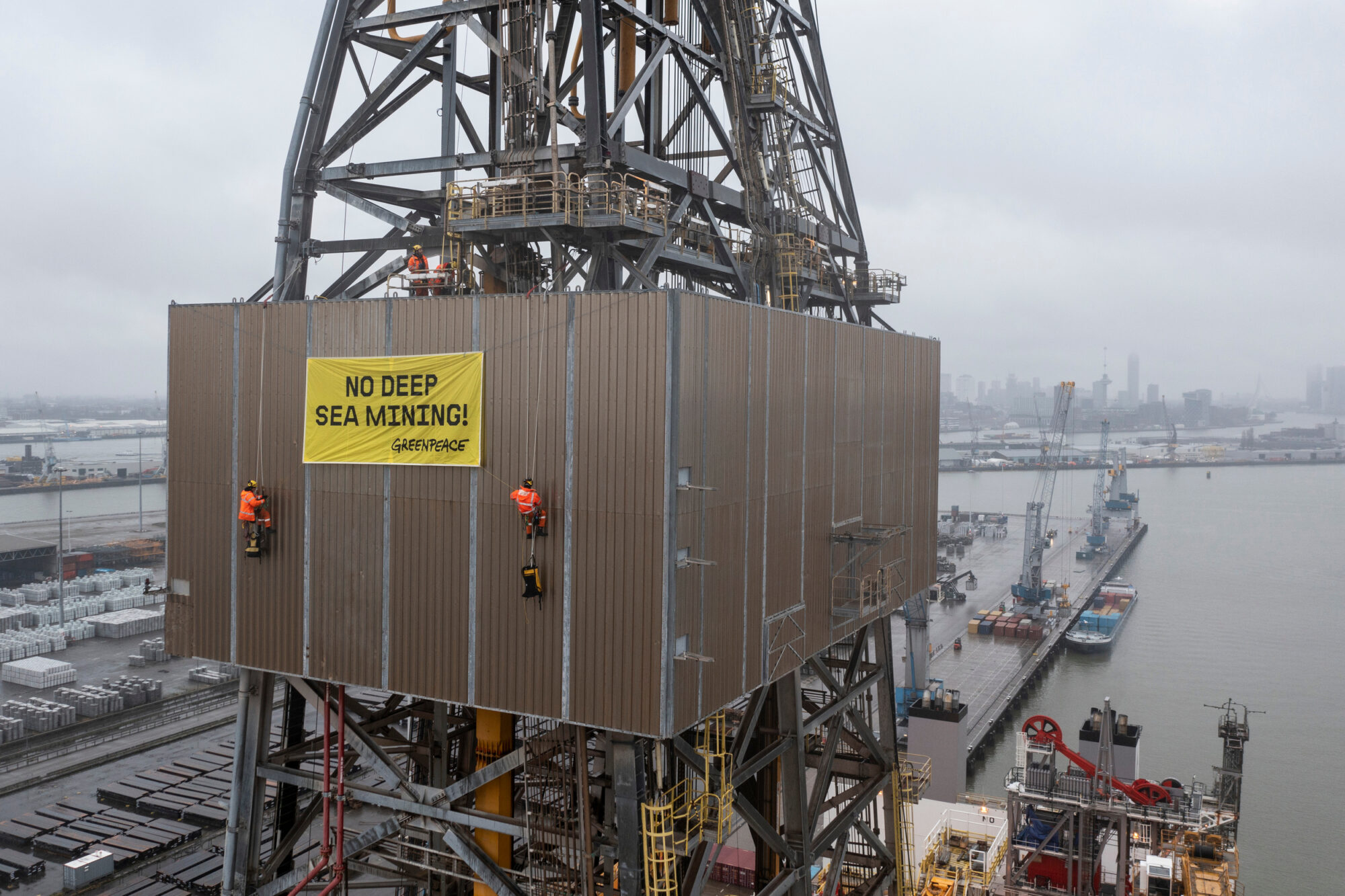 Two climbers hang on the side of a tall tower made of steel beams. Between the climbers is a yellow banner reading "No Deep Sea Mining!". Port infrastructure and open water can be seen in the background.