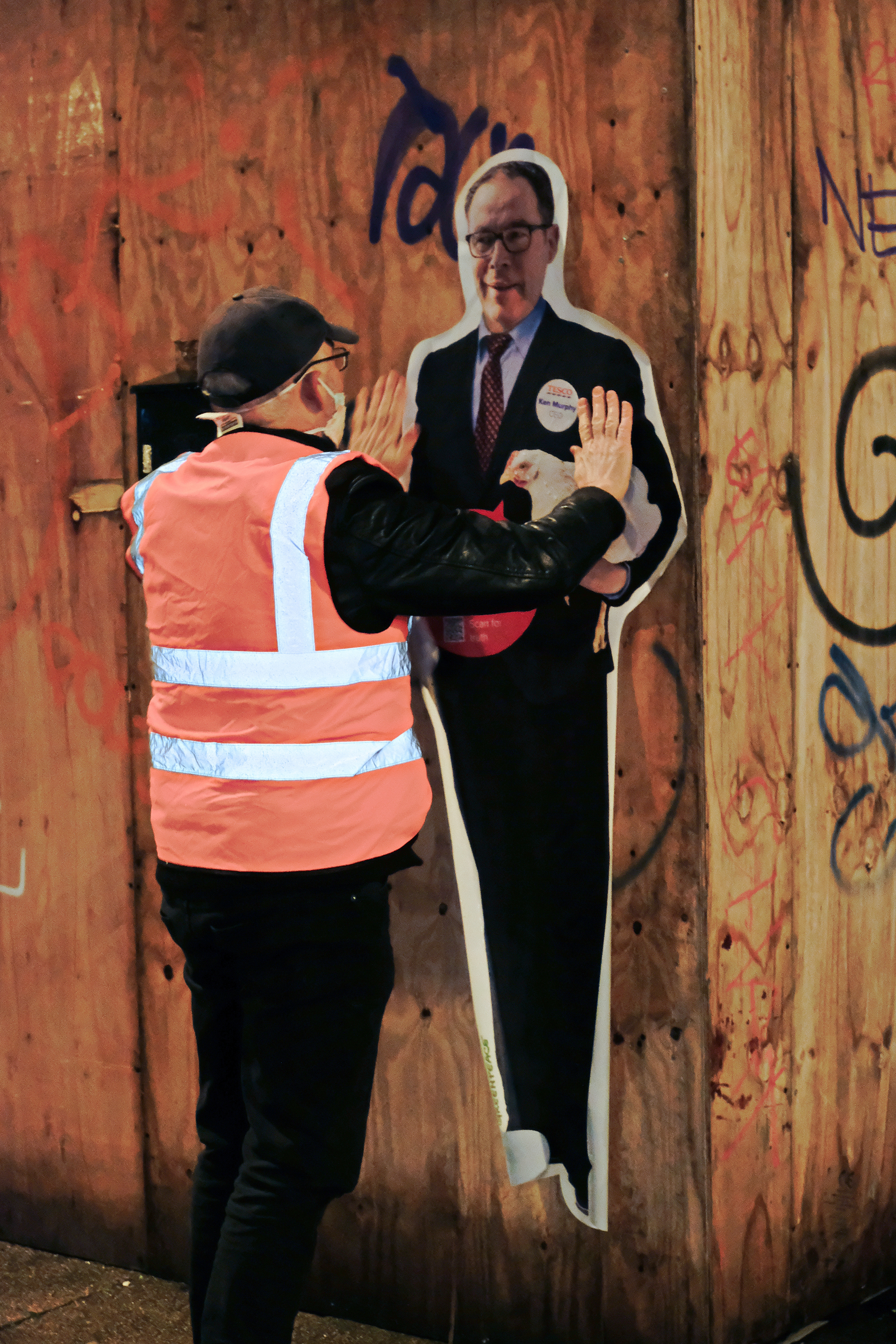 A volunteer putting up a life-size poster depicting Tesco's CEO holding a chicken onto a graffitied wall.