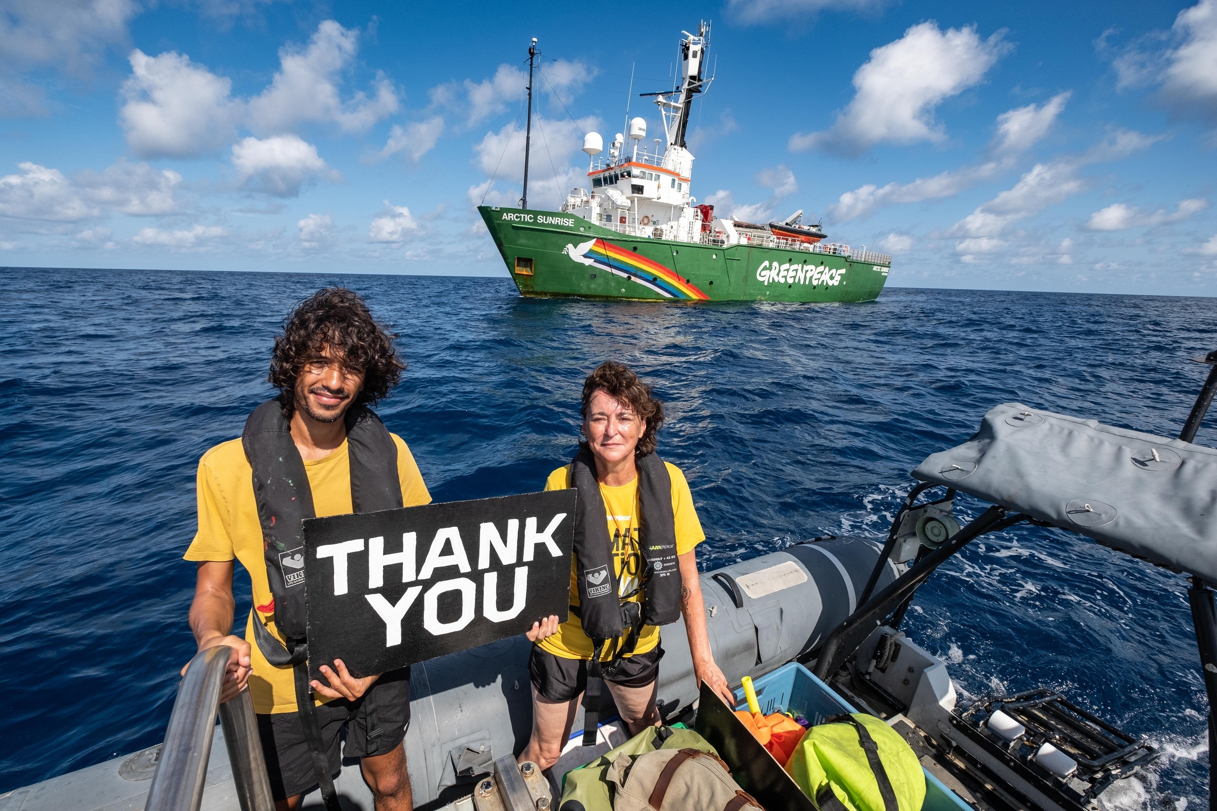 Two people standing on a speedboat in the ocean, holding a sign saying "Thank You". A green ship with "Greenpeace" painted across can be seen behind them.