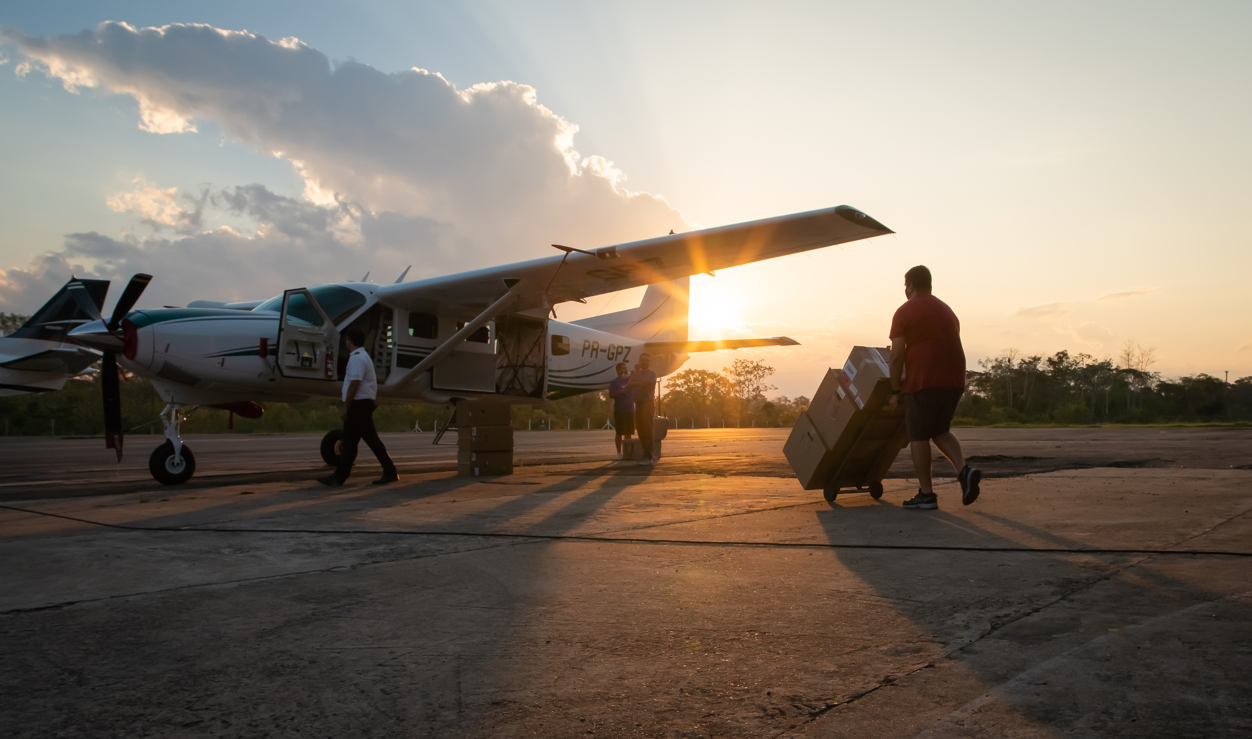 The sun sets behind a small propellor plane, as a person pushes a trolley loaded with large cardboard boxes towards the open side door.