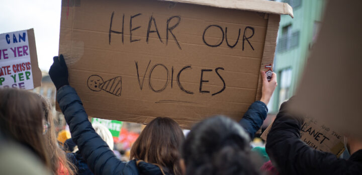 Protester viewed from behind, holding up a handwritten cardboard placard that says 'hear our voices'.