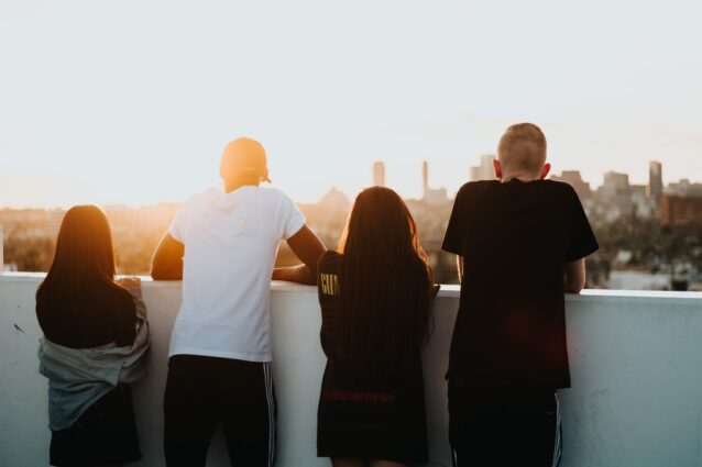 Four people lean on a low wall, facing away from the camera towards a cityscape with the sun setting in the background
