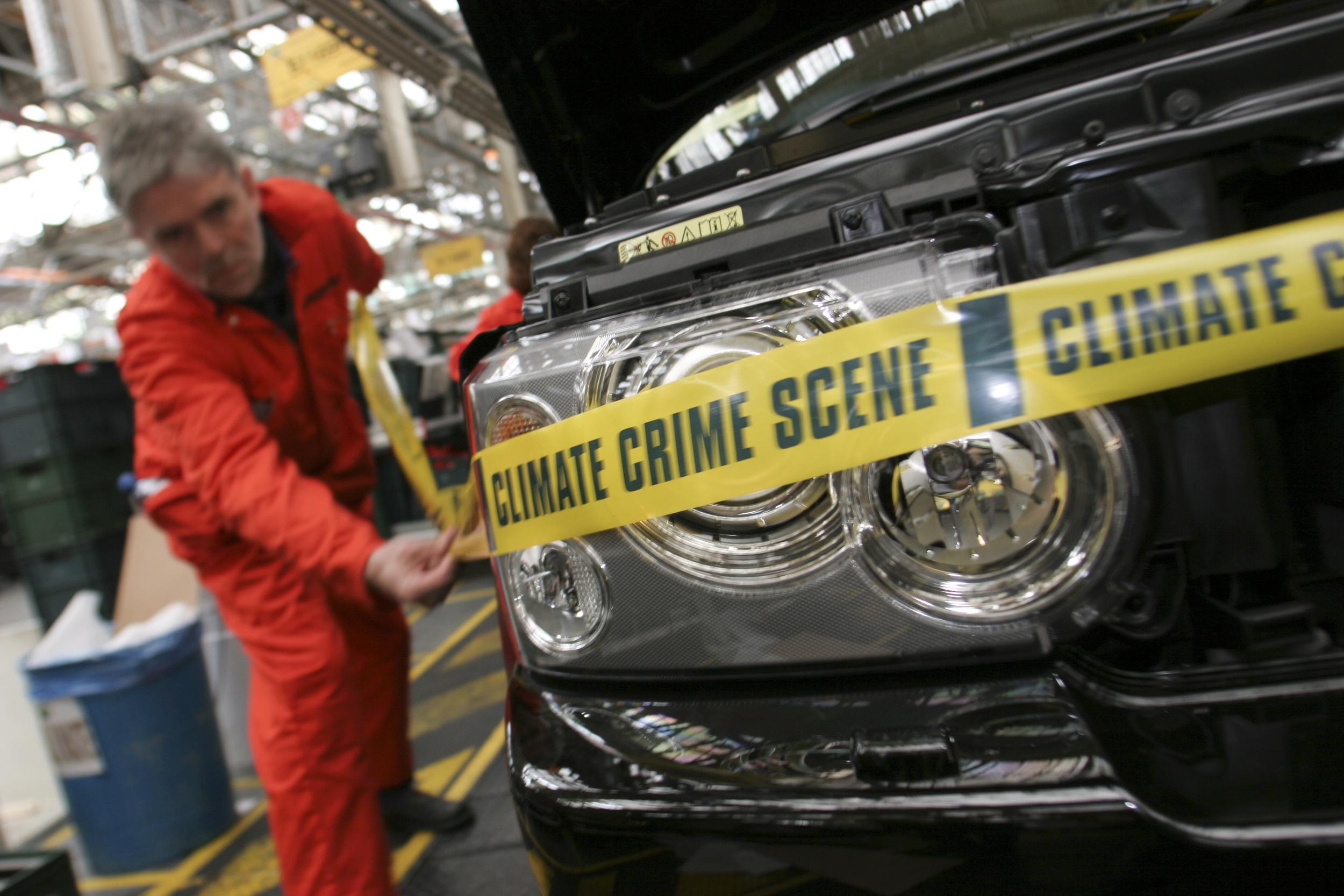 John Sauven, dressed in an orange boiler suit, attaches yellow 'climate crime scene' tape to the front of a car as it moves along a factory assembly line.